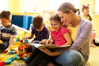 Bild vergr&ouml;&szlig;ern: Happy little girl sitting in teacher's lap in kindergarten and reading a book while the other children are playing. (Happy little girl sitting in teacher's lap in kindergarten and reading a book while the other children are playing., ASCII, 116 compon